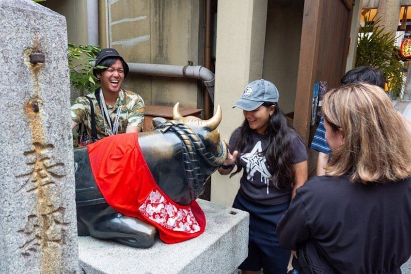 Una estatua de toro con un pañuelo rojo, gente interactuando con ella y un pilar de piedra con esculturas. 4