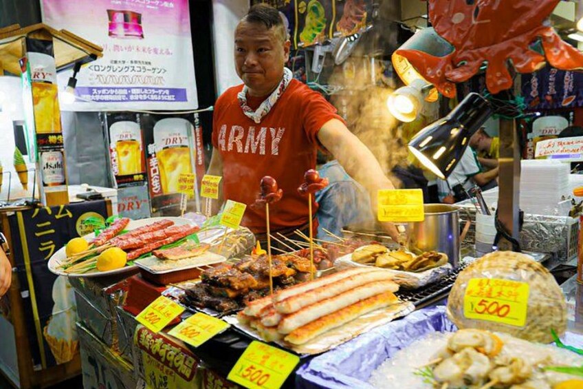 En gatuförsäljare som säljer olika sorters grillat kött och spett, med en skylt som visar priserna och en röd t-shirt med texten ”ARMY”. 1