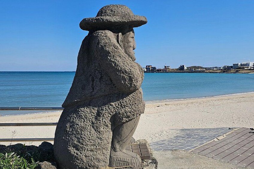 A stone statue of a figure with a hat, a beach, and a clear blue sky. 3
