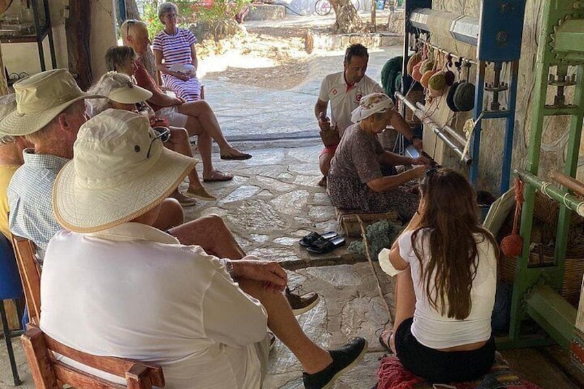 A group of people sitting on chairs in a shaded outdoor area, with a woman knitting. 1