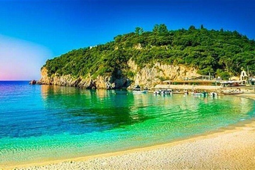 A coastal landscape with a clear blue sea, a sandy beach, and a green hill with buildings. 5