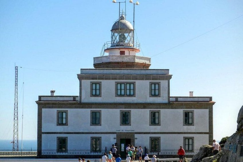 A coastal lighthouse with a weather station on top, a clear sky, and a few people near the entrance. 4