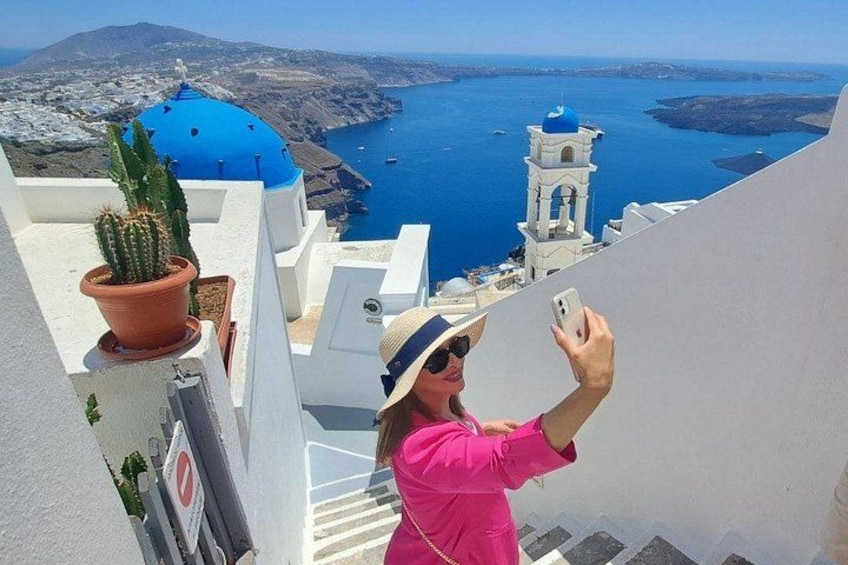 A person taking a selfie on a balcony overlooking a coastal view with a blue-domed building and a white tower. 3