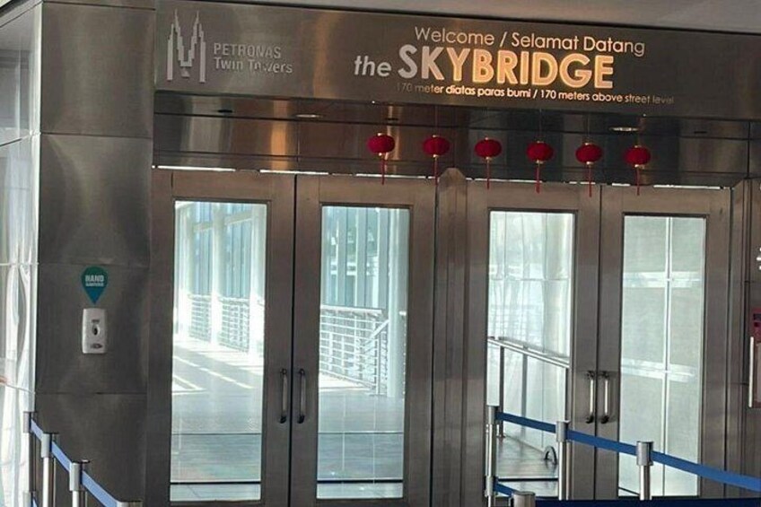 Un ascensore metallico con porte in vetro, un cartello con la scritta 'Welcome / Selamat Datang the SKYBRIDGE' e lanterne rosse sopra. 5
