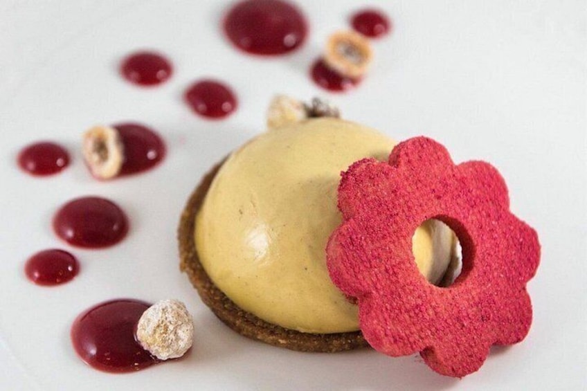 Een dessert met een romige, gele bovenlaag, een rode decoratie in de vorm van een bloem, en verspreide rode bessen en een stukje chocolade op een wit bord. 5