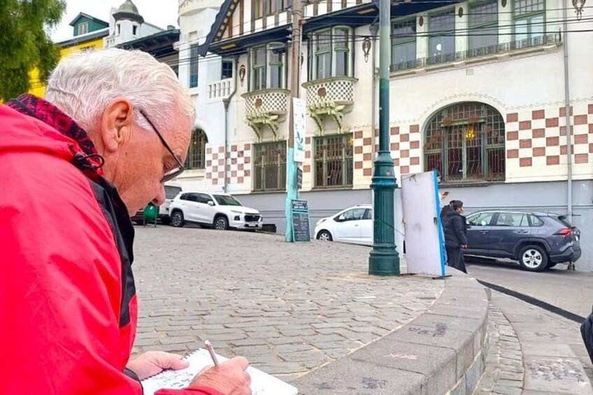 Une rue pavée où l’on voit une personne vêtue d’une veste rouge en train d’écrire sur une feuille de papier, une voiture blanche garée sur le bord et, au fond, un bâtiment à la façade à carreaux. 2
