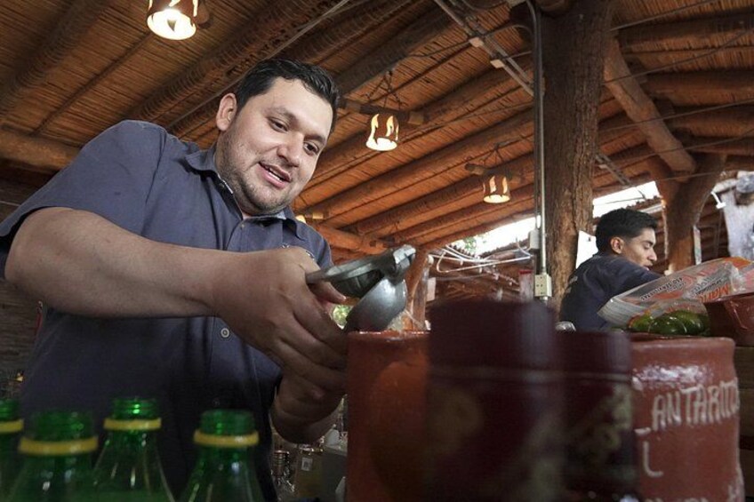 A person pouring a liquid into a container, with bottles and a wooden structure in the background. 3