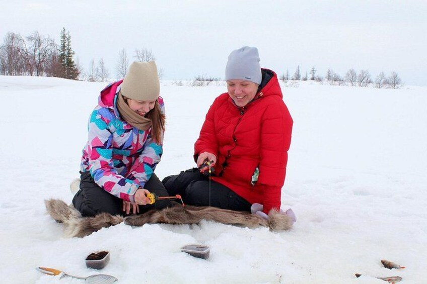 Two people sitting on the snow, one in a colorful jacket and the other in a red jacket, with a log and some tools in front of them. 1