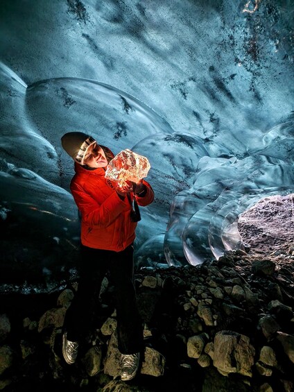 A person in a red jacket holding a crystal, standing in front of a glacier. 4