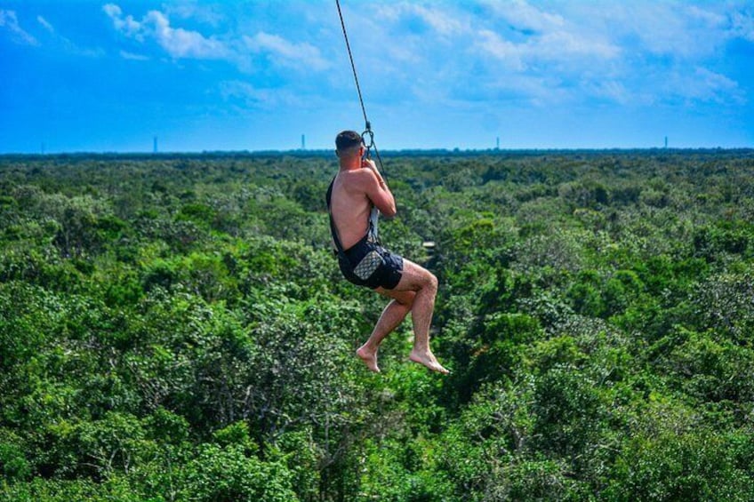 Una persona sospesa a mezz'aria da un cavo, indossa pantaloncini da bagno, con sullo sfondo una vegetazione verdeggiante e un cielo sereno. 4