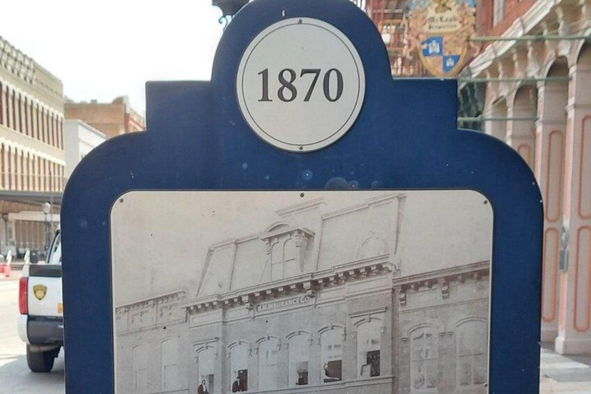 En historisk minnesplatta med siffran 1870, en illustration av en byggnad och en gatubild med fordon. 2