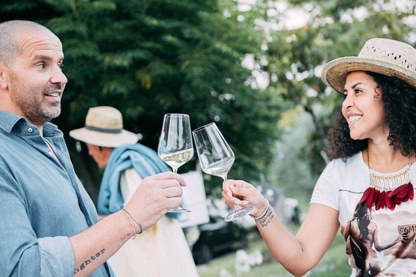 Two people holding wine glasses, one with a tattoo on their arm, in an outdoor setting with trees in the background. 3