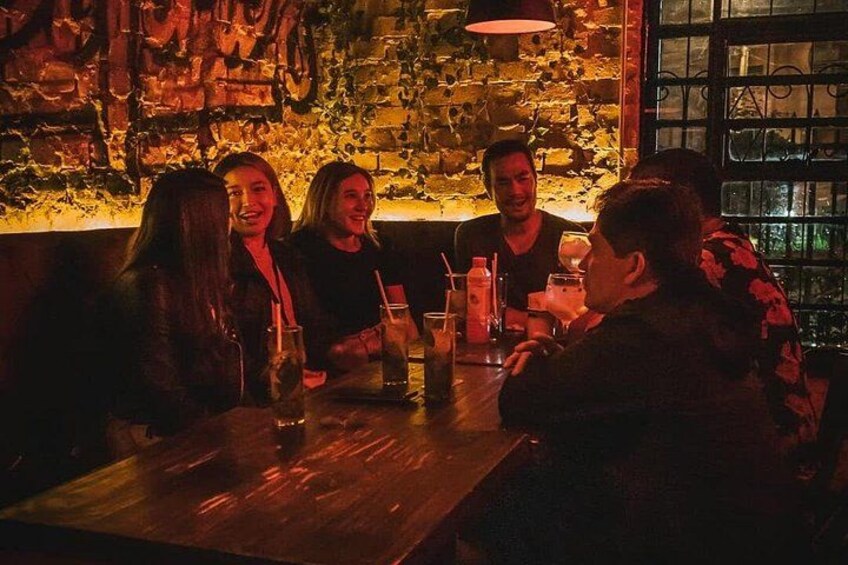 Een groep mensen zit aan een tafel in een schemerig verlicht restaurant, met een bakstenen muur en een raam op de achtergrond. 2