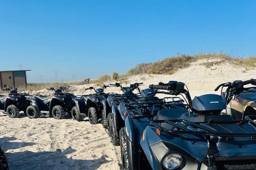 En rad fyrhjulingar står parkerade på en sandstrand med sanddyner i bakgrunden. 5