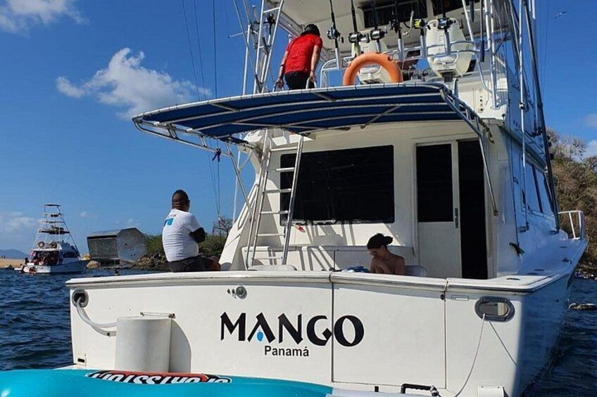 A boat named 'MANGO Panama' with two people on deck, another boat in the background, and calm water. 4