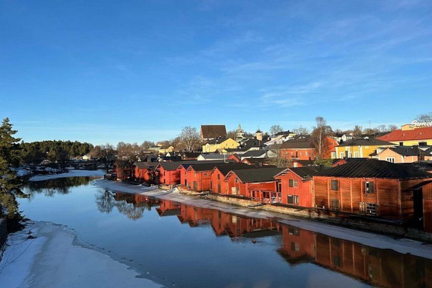 A calm river reflecting red buildings, a clear sky, and a distant town. 1