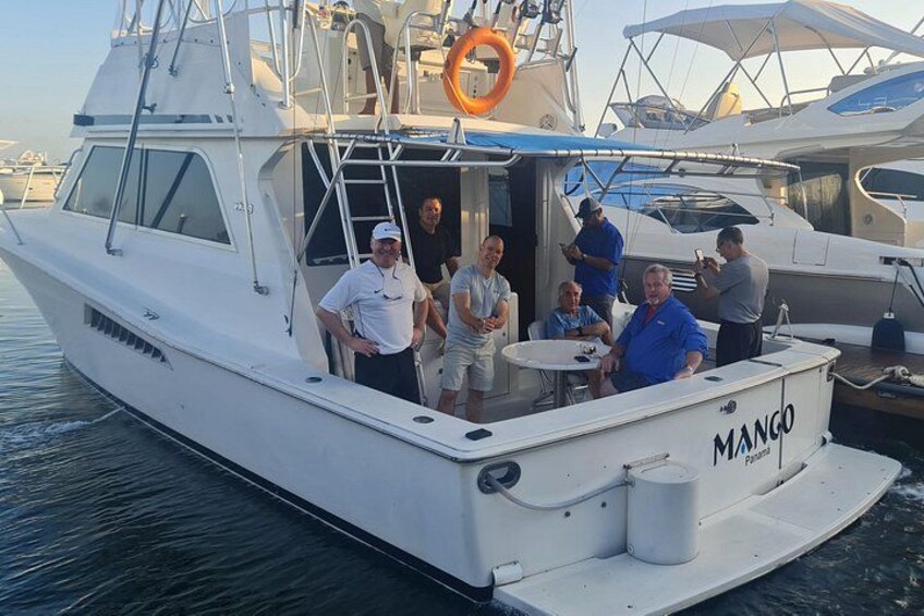 A white boat named 'Mango' with people on board, docked at a marina with other boats. 1