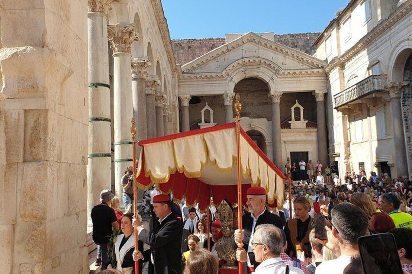 Un edificio histórico con columnas y arcos, una multitud de personas y una procesión ceremonial con toldo rojo y blanco. 5