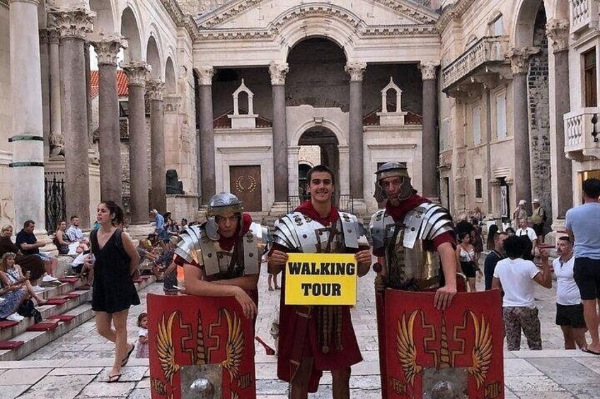 A historical building with columns and arches, a group of people in Roman armor, and a sign that reads 'WALKING TOUR'. 3