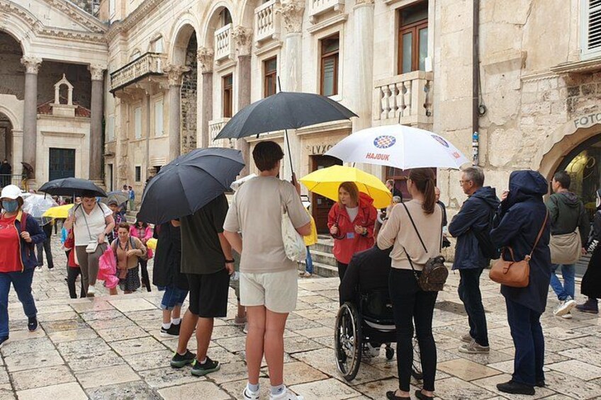 A crowded outdoor area with people walking and standing, some holding umbrellas. 1