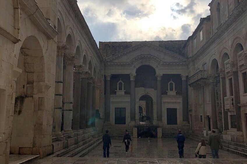 A courtyard with arches and columns, a central building with a pediment, and people walking. 4