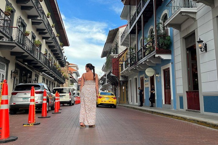 A woman in a white dress walking down a street lined with colorful buildings, traffic cones, and a yellow taxi. 5