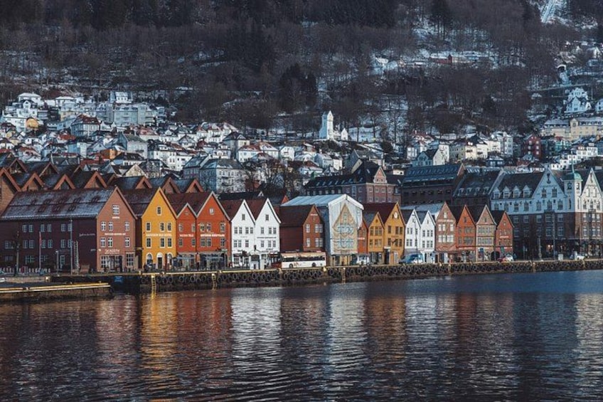 En promenade med farverige huse, der spejler sig i vandet, en bro og en bakket by i baggrunden. 5