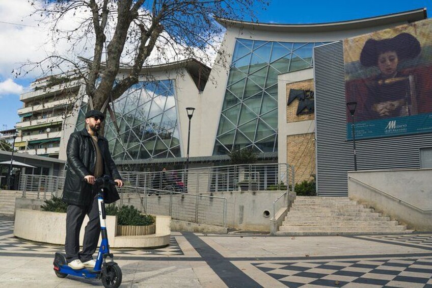 A person on a blue electric scooter in front of a modern building with large glass windows and a checkered pavement. 2