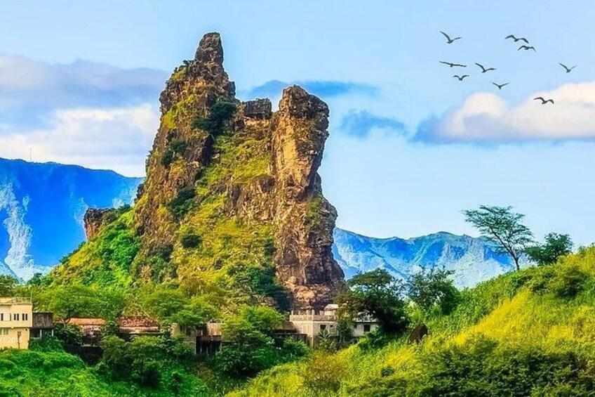 A rocky mountain with a prominent peak, lush green vegetation, and a few buildings at its base. 4
