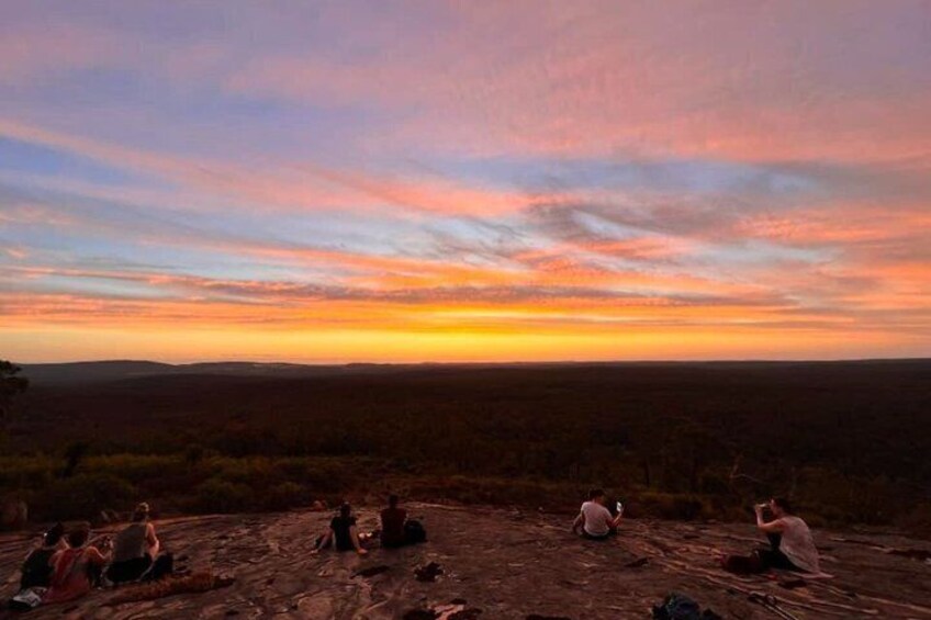 Ein Sonnenuntergang über einer ebenen Landschaft, auf einem Felsen sitzen Menschen. 3