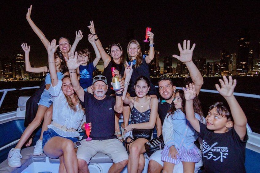 A group of people on a boat with their hands raised, a city skyline in the background, and a body of water. 3