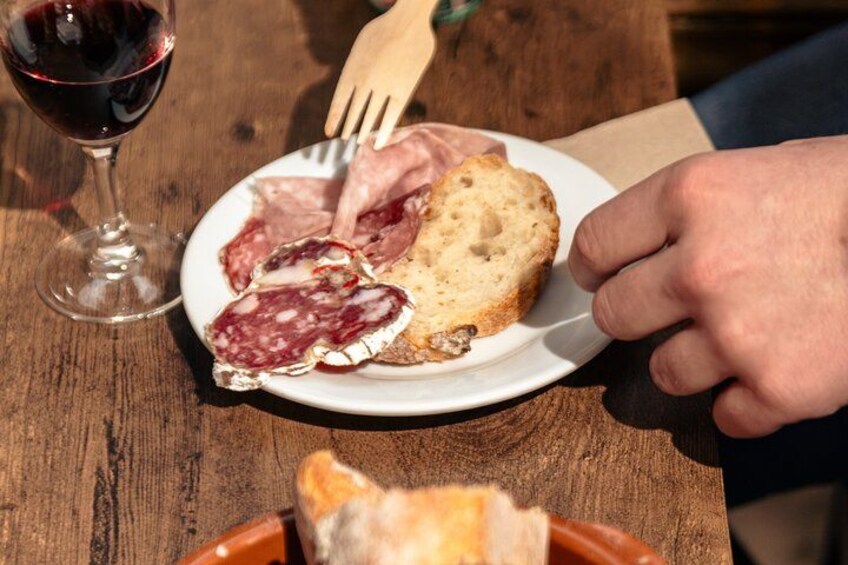 En tallrik med olika sorters kött och bröd, ett glas rött vin och en trägaffel på ett träbord. 5