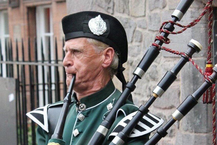 A person playing bagpipes, dressed in a green uniform with a white emblem, standing outdoors with a stone wall and metal fence in the background. 2