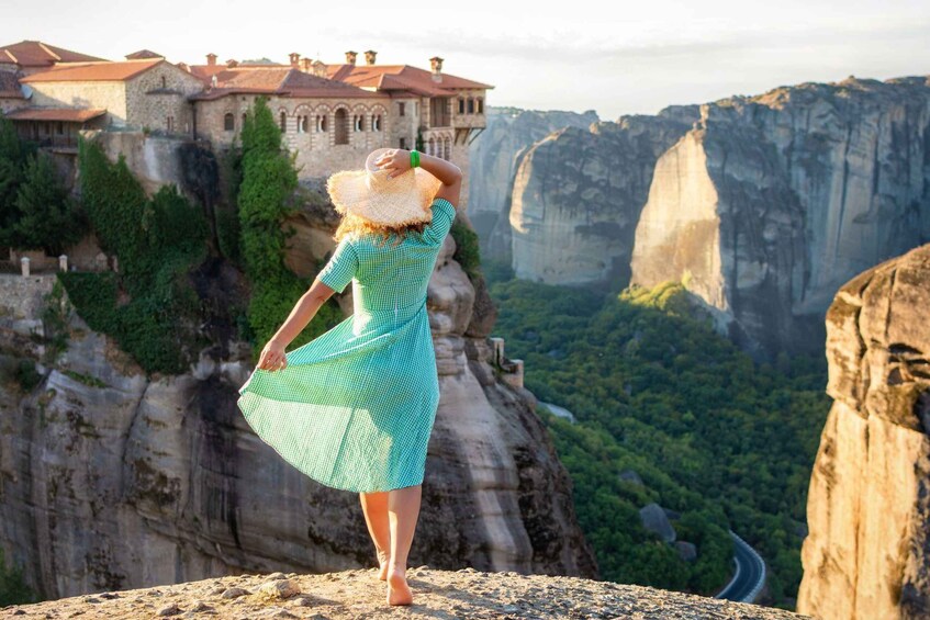 A person standing on a rocky outcrop overlooking a cliff with a building on top and a lush valley below. 1
