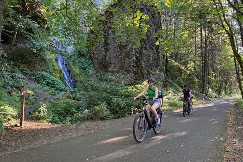Two cyclists on a paved path surrounded by lush greenery and a waterfall. 1