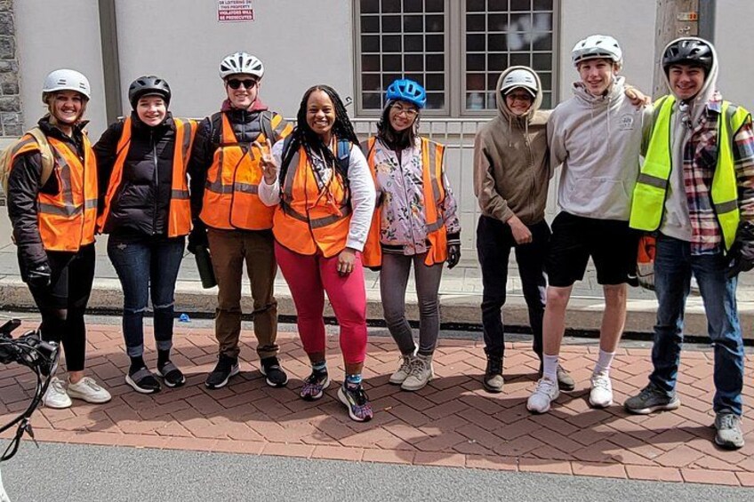 A group of people wearing safety vests and helmets standing on a brick pavement. 4