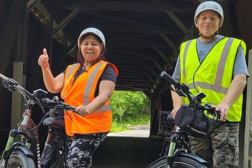 Two people wearing safety vests and helmets, standing next to bicycles in a covered area with greenery in the background. 3