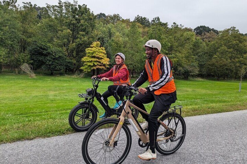 Two people on bicycles, wearing safety vests, on a paved path in a park with trees in the background. 2