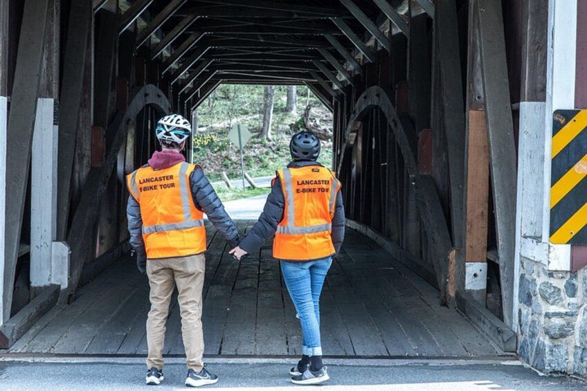 Two people wearing orange vests with 'Lancaster Bike Tour' text, walking under a covered bridge. 5