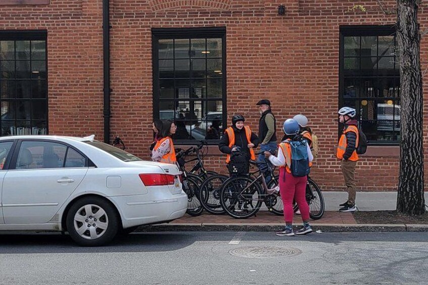 A white car parked on the street, a brick building, and several people with bicycles and safety vests. 2
