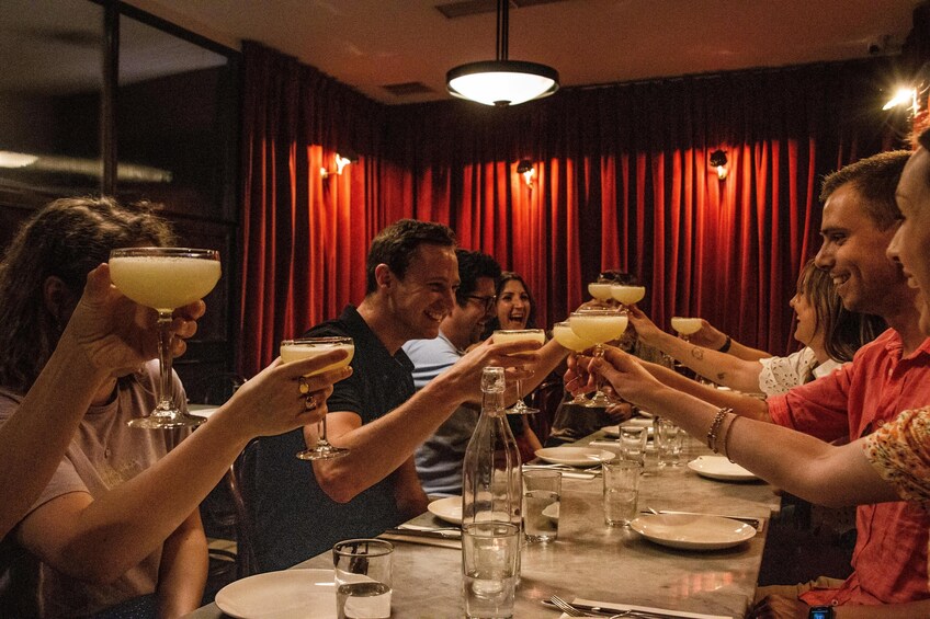 A group of people toasting with drinks at a dinner table in a dimly lit room with red curtains. 1