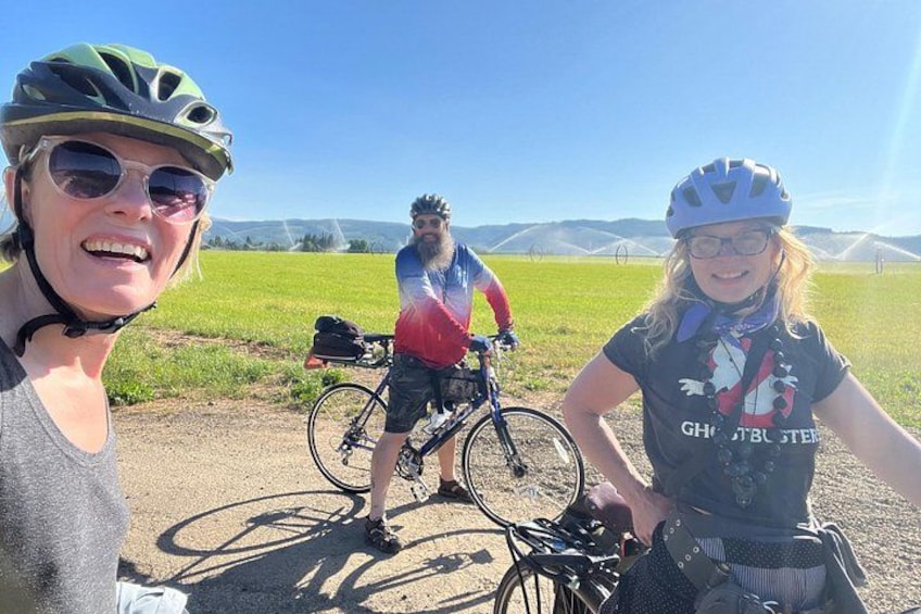 Three people on bicycles in a rural setting with green fields and distant mountains. 4