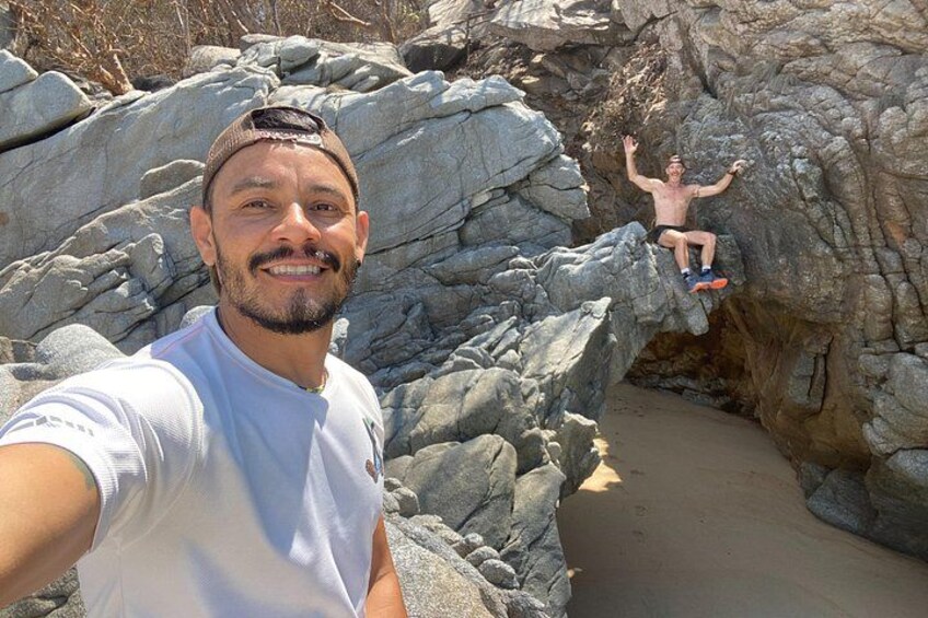 A person taking a selfie with a rocky landscape and another person climbing a rock formation in the background. 1