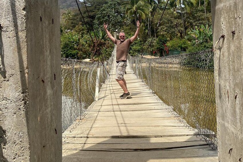A man crossing a suspension bridge over a body of water, surrounded by greenery and a chain-link fence. 2