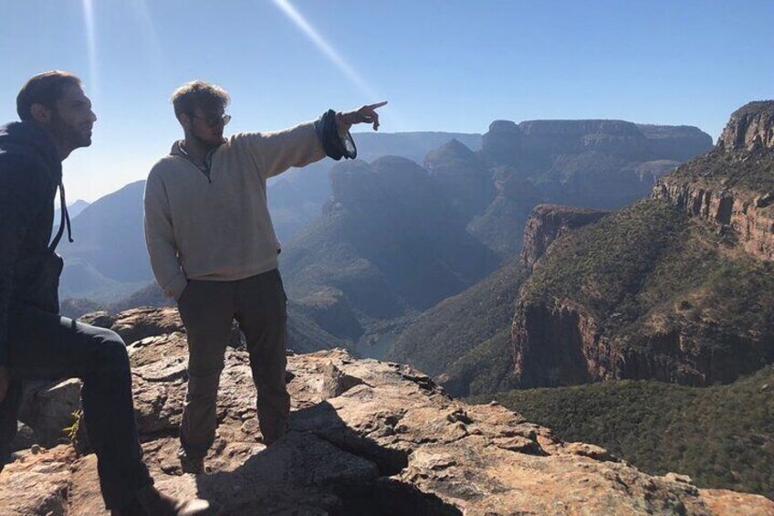 Three people on a rocky outcrop overlooking a canyon with steep cliffs and a clear sky. 1