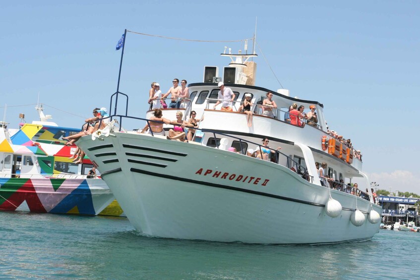 A white boat named 'APHRODITE II' with passengers on deck and a colorful boat nearby. 1
