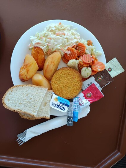 Un plato de comida que incluye ensalada de col, patatas asadas, zanahorias y pan, con salsas y una servilleta sobre una superficie marrón. 3