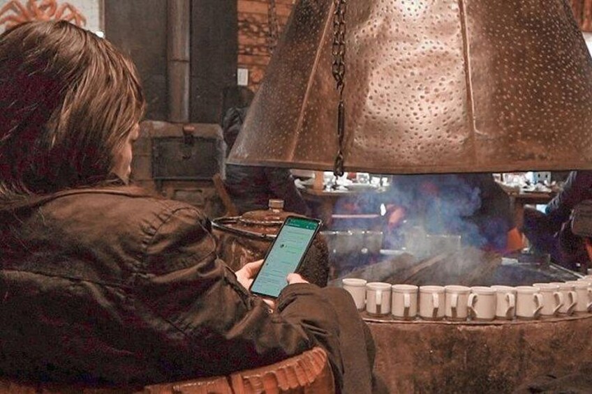 A person sitting by a traditional cooking stove with a metal lid, holding a smartphone. 5