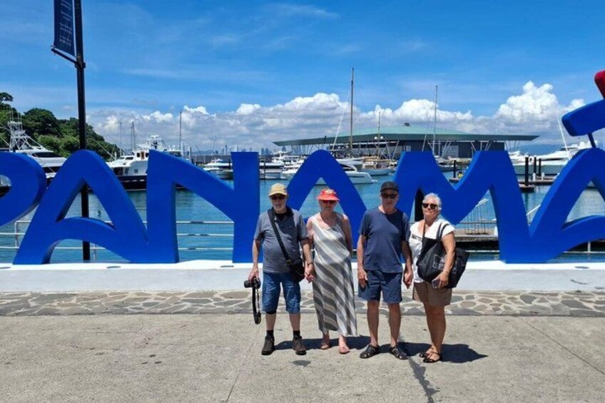 A group of people standing in front of a large blue sign with the word 'PANAMA' on it, with a marina and boats in the background. 3