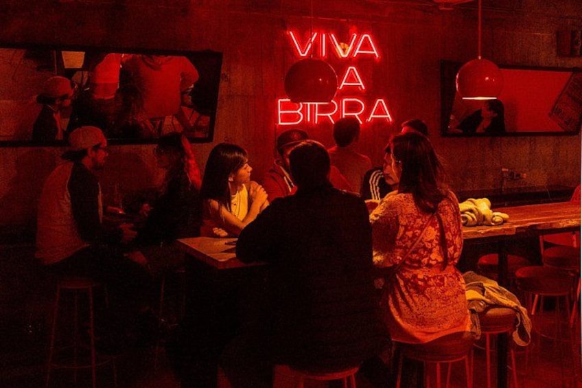 A dimly lit bar with people sitting at tables, a neon sign that reads 'VIVA A BIRRA,' and a mirror on the wall. 2